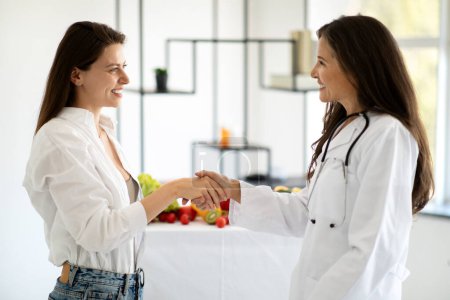 Photo for Cheerful european middle aged lady doctor nutritionist in white coat advises millennial woman patient, shaking hands in clinic interior. Health care, weight loss, professional help - Royalty Free Image