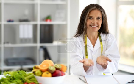 Téléchargez les photos : Heureuse femme européenne d'âge moyen médecin nutritionniste tenir l'espace libre à table avec des fruits, des légumes à l'intérieur du bureau de la clinique. Travail, profession professionnelle et soins de santé, annonce, offre - en image libre de droit