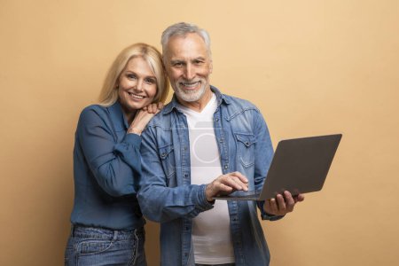Téléchargez les photos : Aimer joyeux conjoints âgés attrayants heureux mari marié et femme en utilisant ordinateur portable PC sur fond beige, regarder beau film en ligne, websurf, défilement sur Internet - en image libre de droit