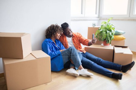 Photo for Happy black couple moving to new home and taking selfie among cardboard boxes, loving young african american spouses making photo in their apartment, celebrating relocation, free space - Royalty Free Image