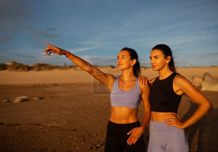 Téléchargez les photos : Souriant assez mince milléniaux jumeaux caucasiens sœurs femmes pointant du doigt à la plage de la mer. Entraînement matinal, sports en plein air, soins de santé, publicité et offre, fitness en été - en image libre de droit