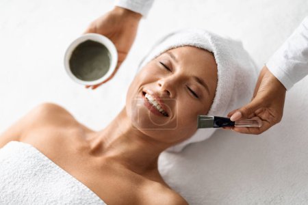 Photo for Beauty Treatments. Cosmetologist applying clay mask on face of middle aged woman at luxury spa salon, relaxed mature female enjoying wellness day at beauty center, top view shot, closeup - Royalty Free Image