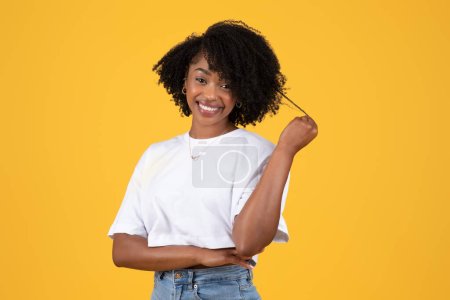 Foto de Mujer rizada negra joven positiva en camiseta blanca toca el cabello, coquetea, disfruta del tiempo libre, aislado sobre fondo naranja, estudio. Moda, estilo de vida, anuncio y oferta, emociones humanas - Imagen libre de derechos