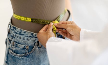 Photo for Mature caucasian lady doctor nutritionist in white coat measures waist of patient with measuring tape in office interior, cropped, close up. Medical examination, weight loss and diet - Royalty Free Image