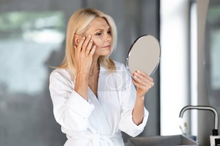 Photo for Skin Aging. Attractive Mature Woman Holding Mirror And Looking At Her Wrinkles Near Eyes, Beautiful Senior Female Wearing Bathrobe Checking Face, Noticing Age Changes, Standing In Bathroom Interior - Royalty Free Image