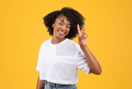 Foto de Joven y alegre dama rizada negra en camiseta blanca haciendo señal de paz con la mano, aislada sobre fondo naranja, estudio. Gesto, estilo de vida, anuncio y oferta, tiempo libre y diversión - Imagen libre de derechos