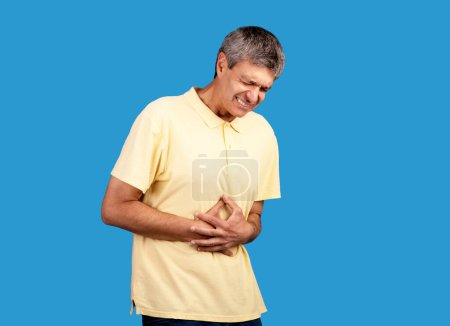 Photo for Abdominal Pain. Mature man suffering from stomachache having painful spasm in belly, touching stomach on blue background, studio shot. Gastritis disease, health problem concept - Royalty Free Image