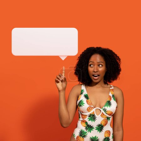 Téléchargez les photos : Émotionnel attrayant millénaire femme afro-américaine en maillot de bain coloré pointant vers blanc ballon de parole blanc, isolé sur fond orange. Dame noire partage des idées sur de belles vacances d'été - en image libre de droit
