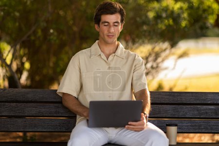 Photo for Handsome young man in casual outfit independent contractor sitting on bench outdoors at public park, using modern pc computer laptop, working or chatting with clients. Remote job, freelance - Royalty Free Image