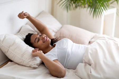 Téléchargez les photos : Portrait de belle femme afro-américaine détendue étirant les mains après le réveil, jeune femme noire se sentant bien, allongée dans un lit confortable à la maison, reposant dans une chambre confortable, gros plan - en image libre de droit