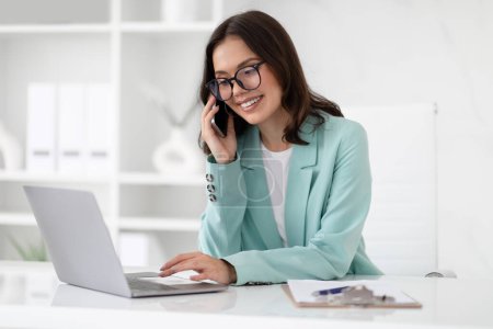 Photo for Cheerful young caucasian businesswoman in suit and glasses typing on computer, enjoys professional occupation, calling by phone in office interior. Business and work, official lifestyle and offer - Royalty Free Image