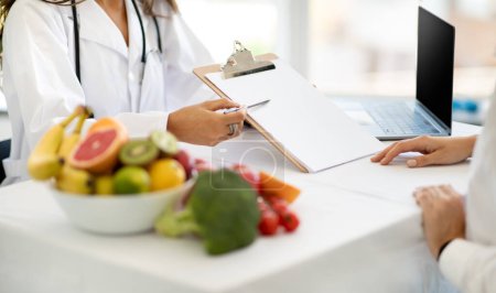 Photo for European middle aged woman doctor nutritionist in white coat show diet program to lady patient in clinic interior, cropped. Proper nutrition, health care, treatment, weight loss with professional - Royalty Free Image