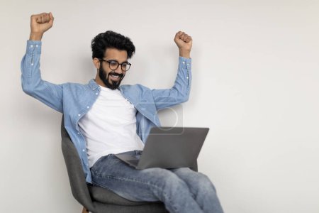 Photo for Online Trading. Happy Young Indian Guy Using Laptop And Celebrating Success, Overjoyed Eastern Man Shaking Fists While Sitting In Chair Over White Wall Background, Cheering Big Profit, Copy Space - Royalty Free Image