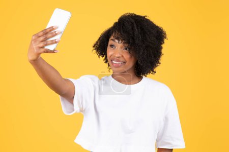 Foto de Alegre joven negro rizado dama en camiseta blanca hace selfie en el teléfono inteligente para las redes sociales, blog, aislado en fondo naranja, estudio. Anuncio y oferta, videollamada, blogger - Imagen libre de derechos