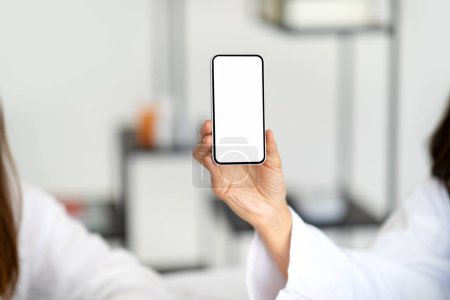 Foto de La nutricionista médica adulta europea en bata blanca recomienda la aplicación por teléfono con pantalla en blanco para el paciente en el interior de la clínica. Gadget para el cuidado de la salud, tratamiento y adelgazamiento - Imagen libre de derechos