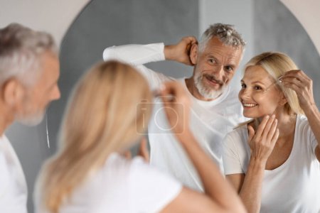 Photo for Family Skincare. Mature Spouses Making Daily Beauty Routine In Bathroom, Happy Senior Couple Smiling To Their Reflection In Mirror, Woman Applying Moisturising Anti-Aging Serum, Selective Focus - Royalty Free Image