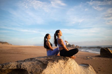 Foto de Alegre milenario gemelos caucásicos hermanas mujeres practican yoga con los ojos cerrados disfrutar de ejercicios de respiración, amanecer en la playa. Entrenamiento matutino, cuidado corporal, deportes juntos y meditación, destello solar - Imagen libre de derechos