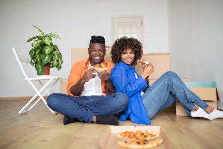 Photo for Lunch Break. Happy Black Spouses Eating Pizza While Celebrating Moving Into New Home, Cheerful Young African American Couple Relaxing Among Cardboard Boxes In Living Room And Smiling At Camera - Royalty Free Image