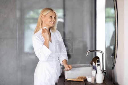Téléchargez les photos : Femme mûre souriante dans la salle de bain nettoyant la peau du cou avec des tampons de coton et regardant dans le miroir, belle femme âgée portant un peignoir blanc rendant la routine quotidienne de beauté à la maison, espace de copie - en image libre de droit