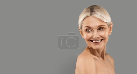 Foto de Anuncio de belleza. Mujer madura sonriente mirando el espacio de copia en el fondo del estudio gris, atractiva rubia mujer mayor girando en el lugar libre para el diseño de publicidad detrás de su espalda, Panorama - Imagen libre de derechos
