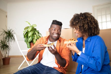 Foto de Cónyuges negros felices comiendo pizza y divirtiéndose en el día de mudanza, alegre pareja afroamericana joven sentada en el piso entre cajas de cartón, almorzando, celebrando la reubicación, primer plano - Imagen libre de derechos