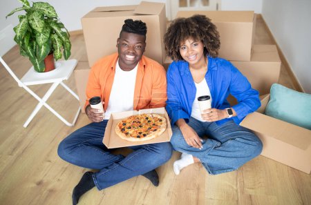 Téléchargez les photos : Déjeuner du jour du déménagement. Joyeux jeune couple noir mangeant de la pizza et buvant du café à emporter tout en étant assis sur le sol parmi des boîtes en carton, joyeux conjoints afro-américains appréciant la nourriture et regardant la caméra - en image libre de droit