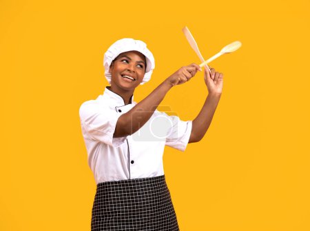 Foto de Mujer Chef Negro alegre divirtiéndose con espátulas en las manos, cocinera afroamericana feliz en uniforme jugando con utensilios de cocina mientras posa aislada en el fondo del estudio amarillo, espacio de copia - Imagen libre de derechos