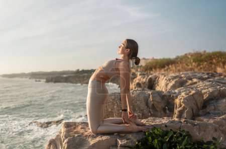 Foto de Calm millennial mujer atleta caucásica en ropa deportiva practica yoga, estirando el cuerpo, disfruta del ejercicio en la playa del mar por la mañana, perfil. Cuidado de la salud, deportes al aire libre, cuidado corporal y estilo de vida - Imagen libre de derechos
