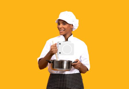 Photo for Culinary Concept. Black Chef Woman Cooking Food In Saucepan Over Yellow Background, Young African American Cook Lady In Uniform Stirring Meal In Stewpot With Spatula And Smiling At Camera, Copy Space - Royalty Free Image
