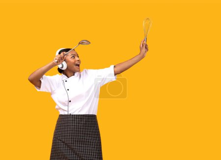 Foto de Chef alegre mujer negra con auriculares inalámbricos bailando con utensilios de cocina en las manos, Happy African American Cook Lady Divirtiéndose, Posando aislado en el fondo del estudio amarillo, Copiar el espacio - Imagen libre de derechos