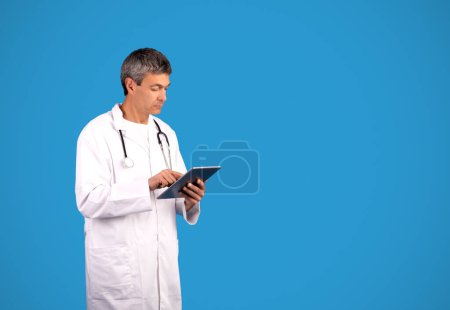 Téléchargez les photos : Médecin homme en manteau blanc à l'aide d'un ordinateur tablette numérique pour le travail posant sur fond bleu en studio. Médecin généraliste avec les cheveux gris debout près de l'espace de copie pour la publicité médicale - en image libre de droit