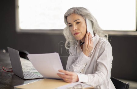 Photo for Business Calls, Phone Communication. Mature Businesswoman Talking On Cellphone Holding Papers, Calling Job Candidates Or Clients Sitting At Workplace Near Laptop In Modern Office Interior - Royalty Free Image