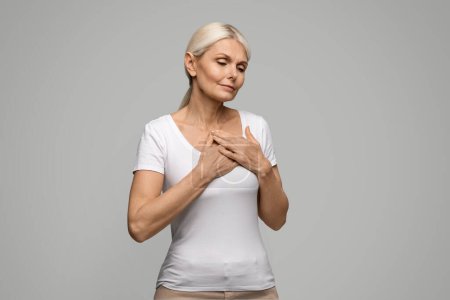 Photo for Heart Attack. Mature blonde woman suffering from severe chest pain, senior female feeling unwell, facing signs of myocardial infarction disease, standing on grey studio background, copy space - Royalty Free Image