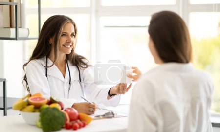 Photo for Cheerful mature caucasian lady doctor nutritionist in white coat advises young woman jar of vitamin pills at table with organic vegetables and fruits in office interior. Health care and diet - Royalty Free Image