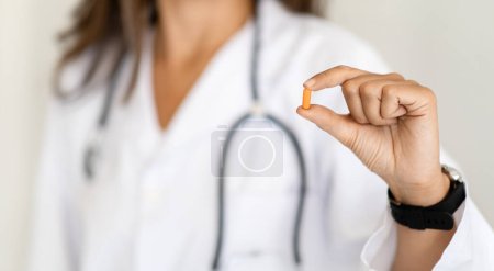 Téléchargez les photos : Femme caucasienne mature médecin en manteau blanc montre pilule capsule, vitamines, complément alimentaire en clinique sur fond studio lumière. Recommandation professionnelle pour les soins de santé, traitement médical - en image libre de droit