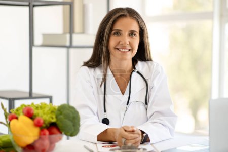 Fröhliche reife kaukasische Ärztin in weißem Mantel am Tisch mit Laptop mit Bio-Früchten im Büroinnenraum. Professionelle Beratung, Gesundheitsempfehlung, Gewichtsverlust, Vitamine
