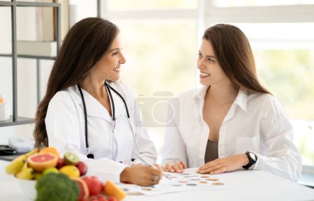 Happy mature caucasian doctor nutritionist in white coat advises young woman at table with organic vegetables and fruits in office interior. Health care, exam, diet plan, professional consultation