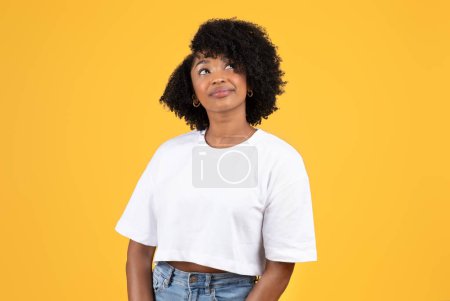 Photo for Positive pretty pensive young black curly lady in white t-shirt looks up at copy space, think, isolated on orange background, studio. Ad and offer, create idea, sale and dreams - Royalty Free Image