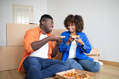 Téléchargez les photos : Jeune couple noir se reposant après avoir déménagé à la maison, assis sur le sol et mangeant de la pizza, joyeux conjoints afro-américains ayant pause déjeuner parmi les boîtes en carton, célébrant la réinstallation dans un nouvel appartement - en image libre de droit