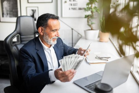 Photo for Positive caucasian senior businessman in suit use phone, computer, hold lots of money dollars in office interior. App, finance work remotely, online business, successful startup and investment - Royalty Free Image