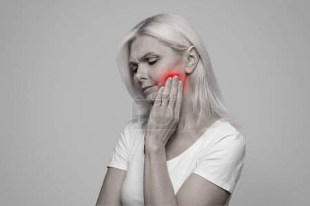 Teeth Problem. Mature Woman Suffering Acute Toothache, Massaging Aching Red Sore Zone, Upset Senior Female Rubbing Jawline, Having Periodontitis, Standing Over Grey Studio Background, BW Shot