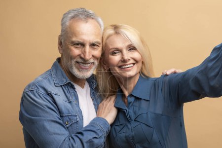Téléchargez les photos : Heureux couple sénior attrayant deux amis âgés homme aux cheveux gris femme blonde en denim vêtements décontractés étreignant, prenant selfie ensemble, isolé sur fond de couleur beige, portrait studio - en image libre de droit