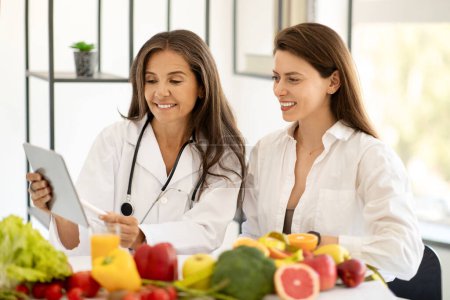 Glücklich reifen kaukasischen Arzt Ernährungsberater in weißem Kittel Beratung junge Frau, zeigen Plan Ernährung auf Tablette am Tisch mit Bio-Gemüse und Obst im Büro Interieur. Gesundheitsfürsorge, Gewichtsverlust