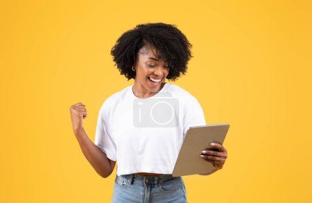 Photo for Cheerful excited young black curly lady in white t-shirt looking tablet, making success gesture, celebrating victory, isolated on orange background, studio. Ad and offer, winner and sale - Royalty Free Image