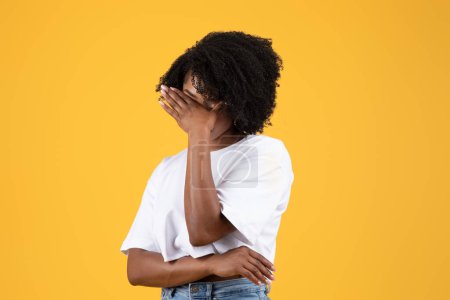 Photo for Sad shy young black curly lady in white t-shirt covers face with hand, isolated on orange background, studio. Embarrassment, bad news reaction, not seeing and human emotions - Royalty Free Image