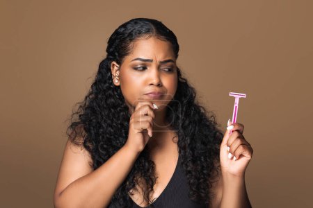 Photo for Wax or shave. Thoughtful latin plus size lady looking at safety razor and touching chin, standing posing isolated on brown studio background - Royalty Free Image