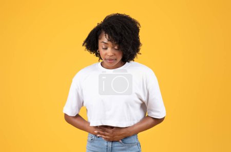 Téléchargez les photos : Malheureuse jeune femme bouclée noire en t-shirt blanc se presse les mains au ventre, souffre de douleurs à l'estomac, isolée sur fond orange, studio. Problèmes de santé, menstruations, empoisonnement, diarrhée - en image libre de droit