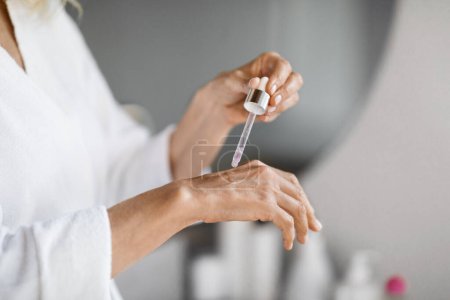 Photo for Mature Woman Applying Moisturusing Serum On Hand With Dropper, Closeup Shot Of Unrecognizable Female In Bathrobe Testing New Beauty Product While Making Selfcare Treatments At Home, Cropped - Royalty Free Image