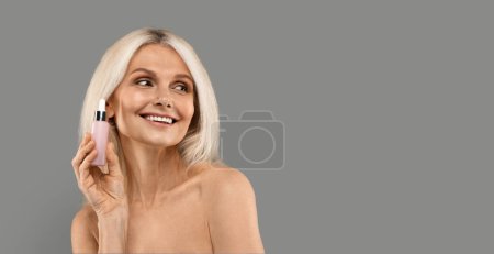 Photo for Anti-Aging Cosmetics. Beautiful Mature Woman Holding Bottle With Face Serum And Looking Aside At Copy Space, Attractive Senior Woman Recomending Beauty Product, Standing On Grey Background, Panorama - Royalty Free Image