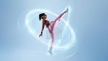 Téléchargez les photos : Sport numérique. Femme noire motivée faisant un coup de pied de karaté, femme sportive levant la jambe, montrant un étirement parfait, femme s'entraînant à l'autodéfense, utilisant un casque VR sur fond de studio bleu, pleine longueur - en image libre de droit